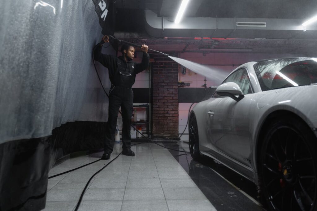 A man uses a pressure washer to clean a white car in an indoor car wash.