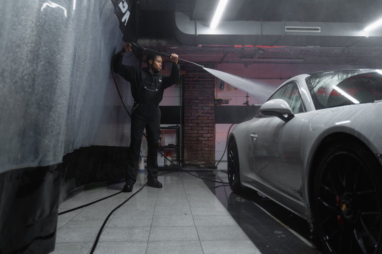 A man uses a pressure washer to clean a white car in an indoor car wash.